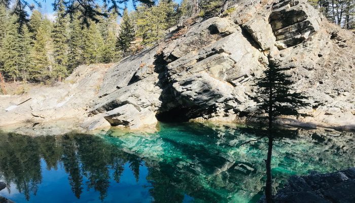 A photo of a beautiful green reflective pool of water in front of an interesting mountain rock with blue sky and trees reflecting in the water. A great place for recovering from stress. Learn how to recover here.